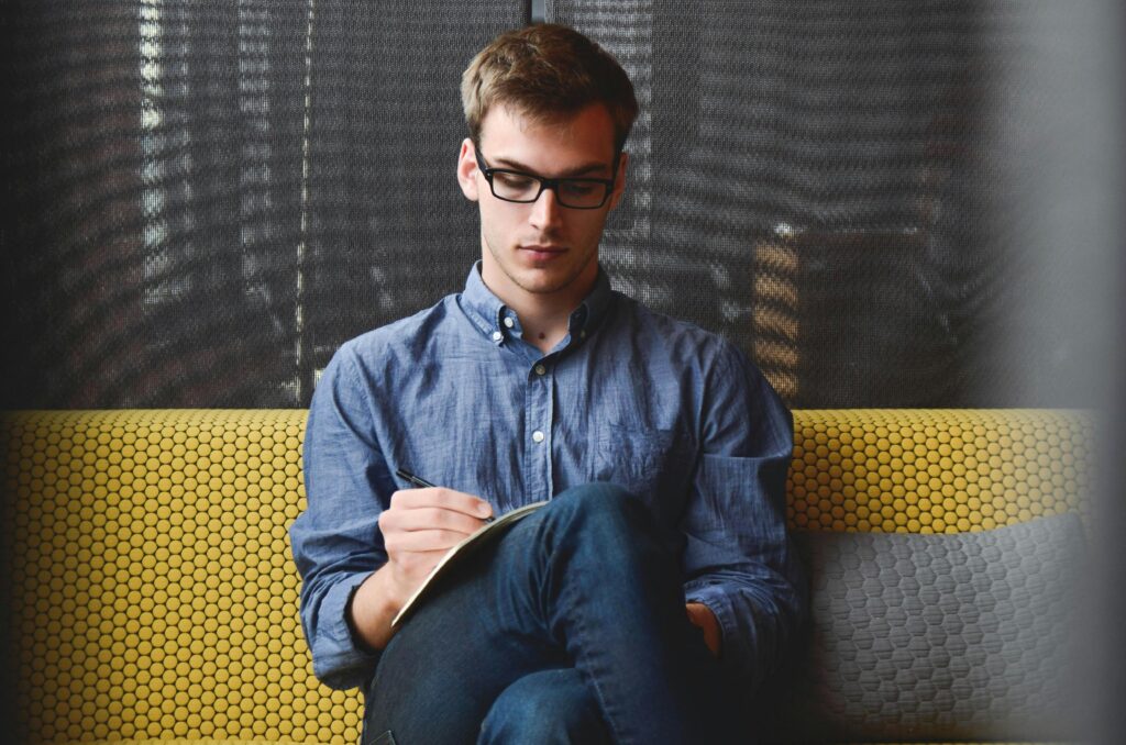 Tarifvergleich und Service A young man in glasses writes in a notebook while sitting on a stylish couch indoors.